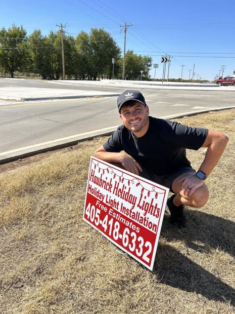 Cameron Haubrick, owner of Haubrick Holiday Lights, beside a company yard sign